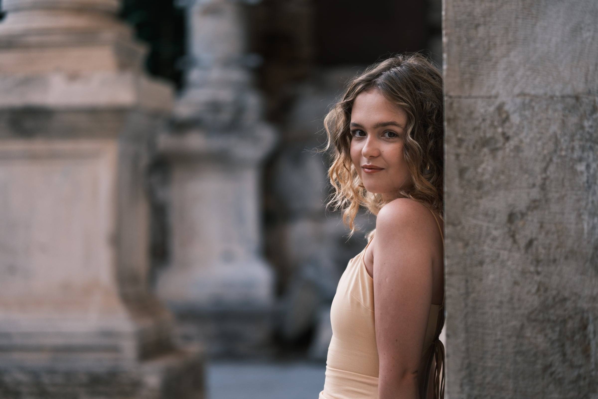 Woman with curly hair looking over her shoulder beside an ancient pillar