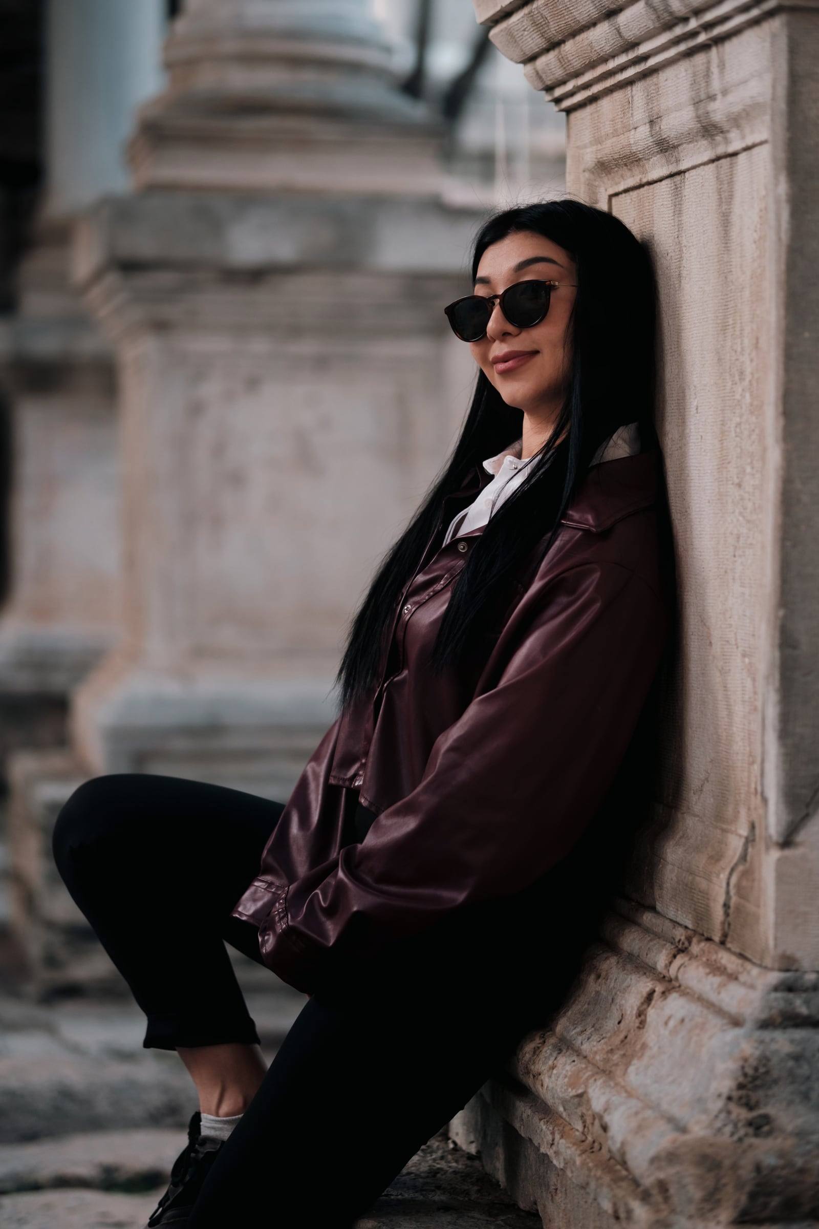 Woman in a leather jacket sitting by an ancient marble wall