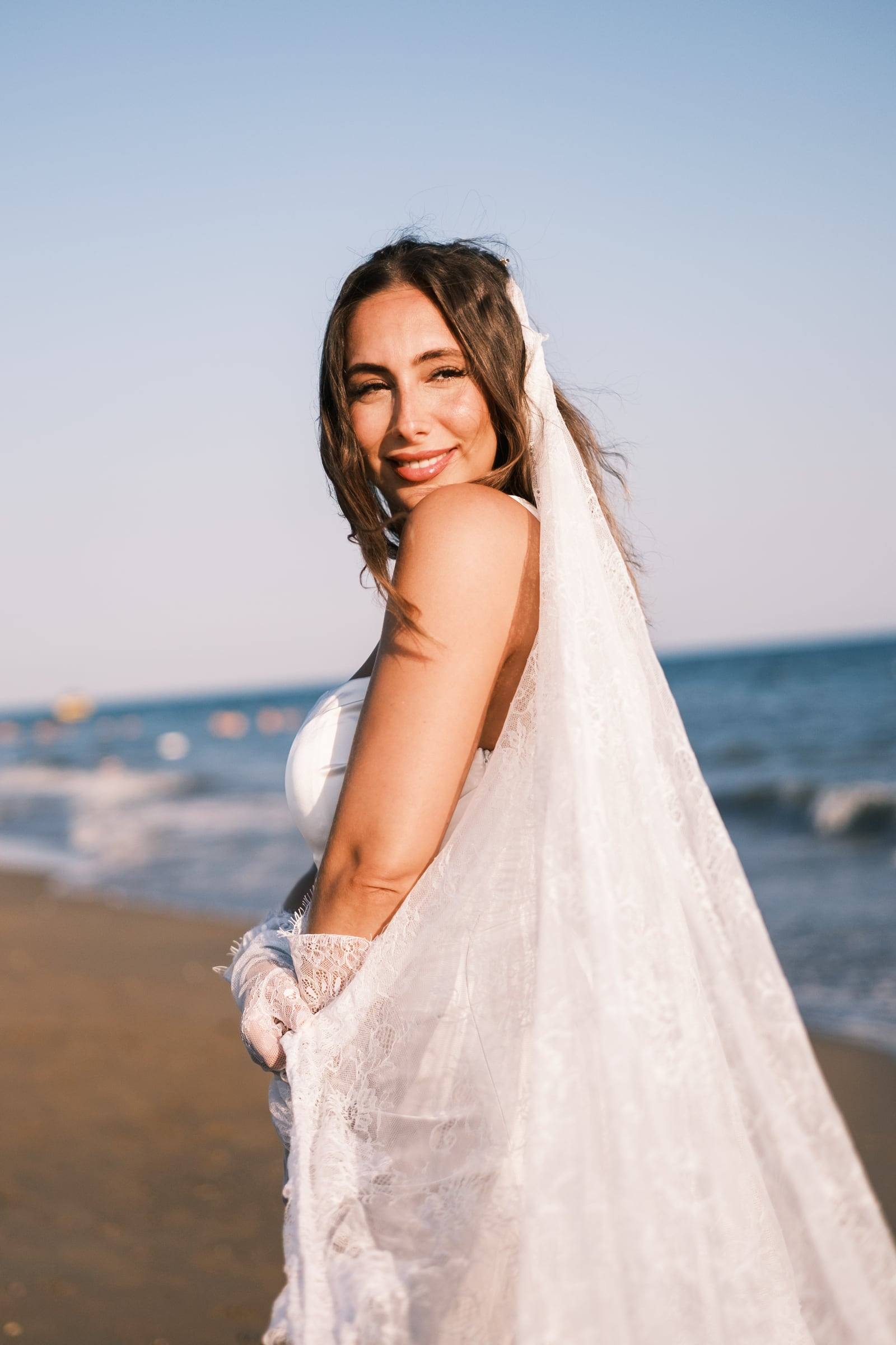 Bride in a lace veil smiling on a sandy beach