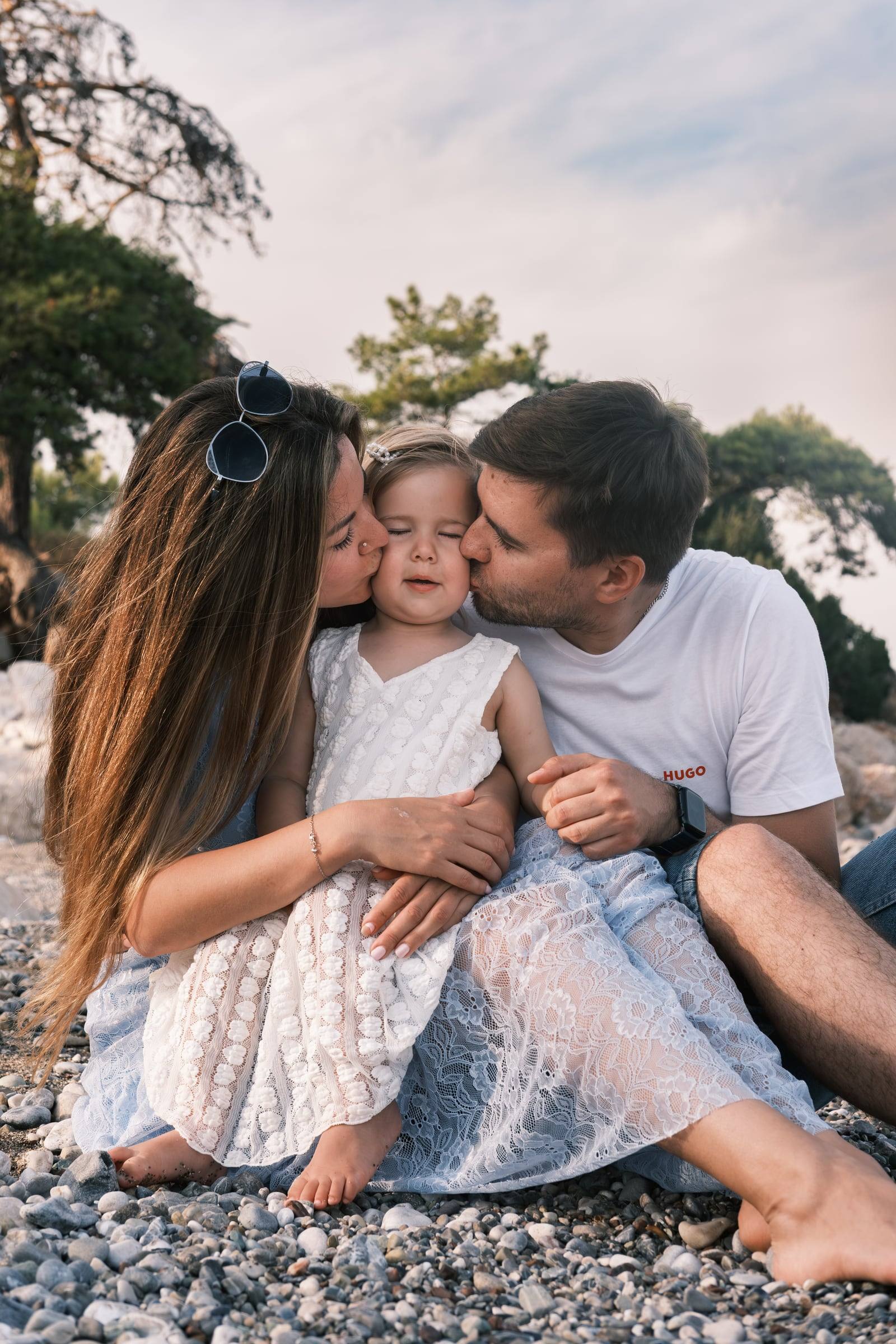 Parents kissing their toddler on a pebble beach under pine trees