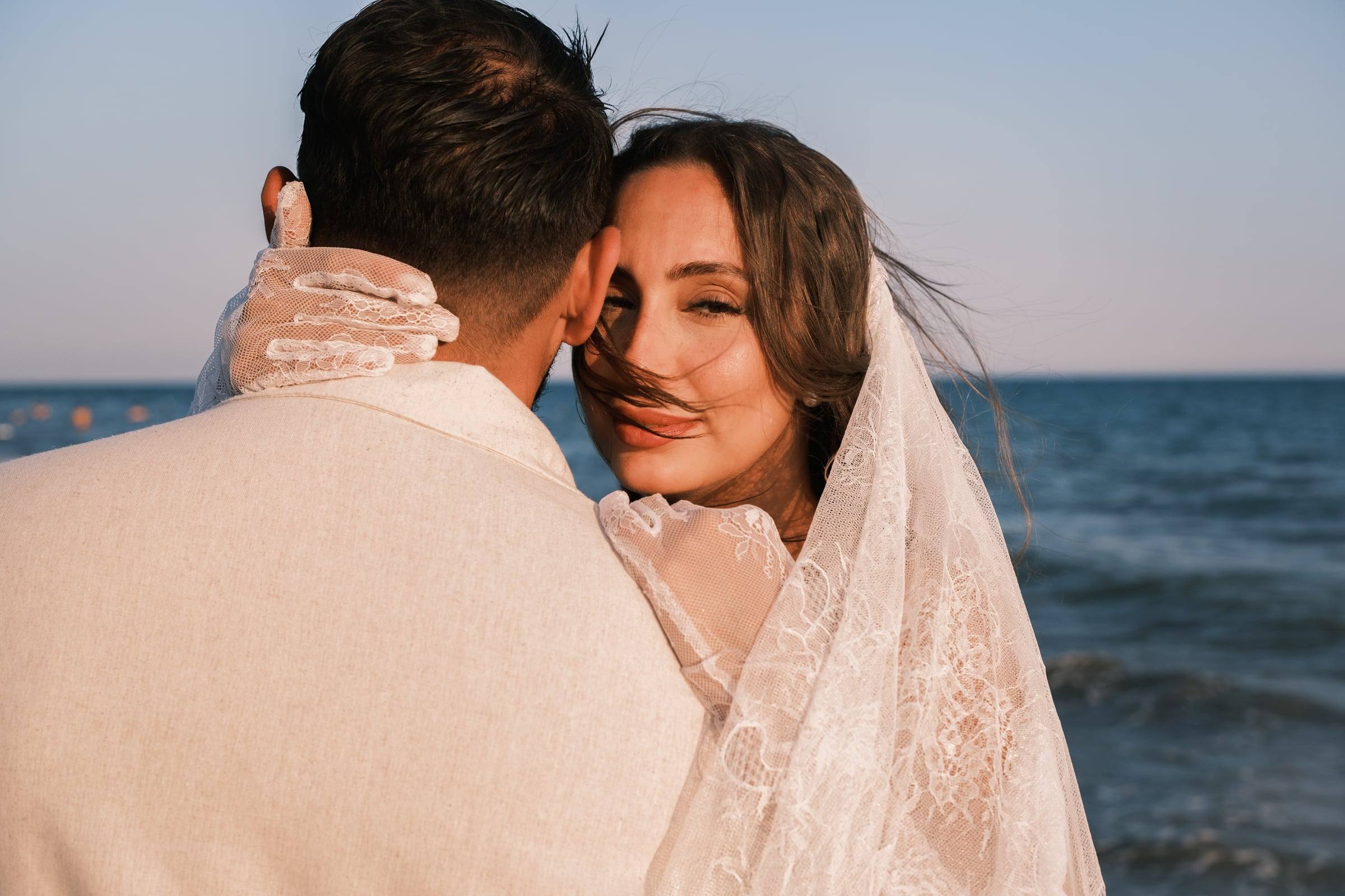 Bride in a veil looking over the groom's shoulder by the sea