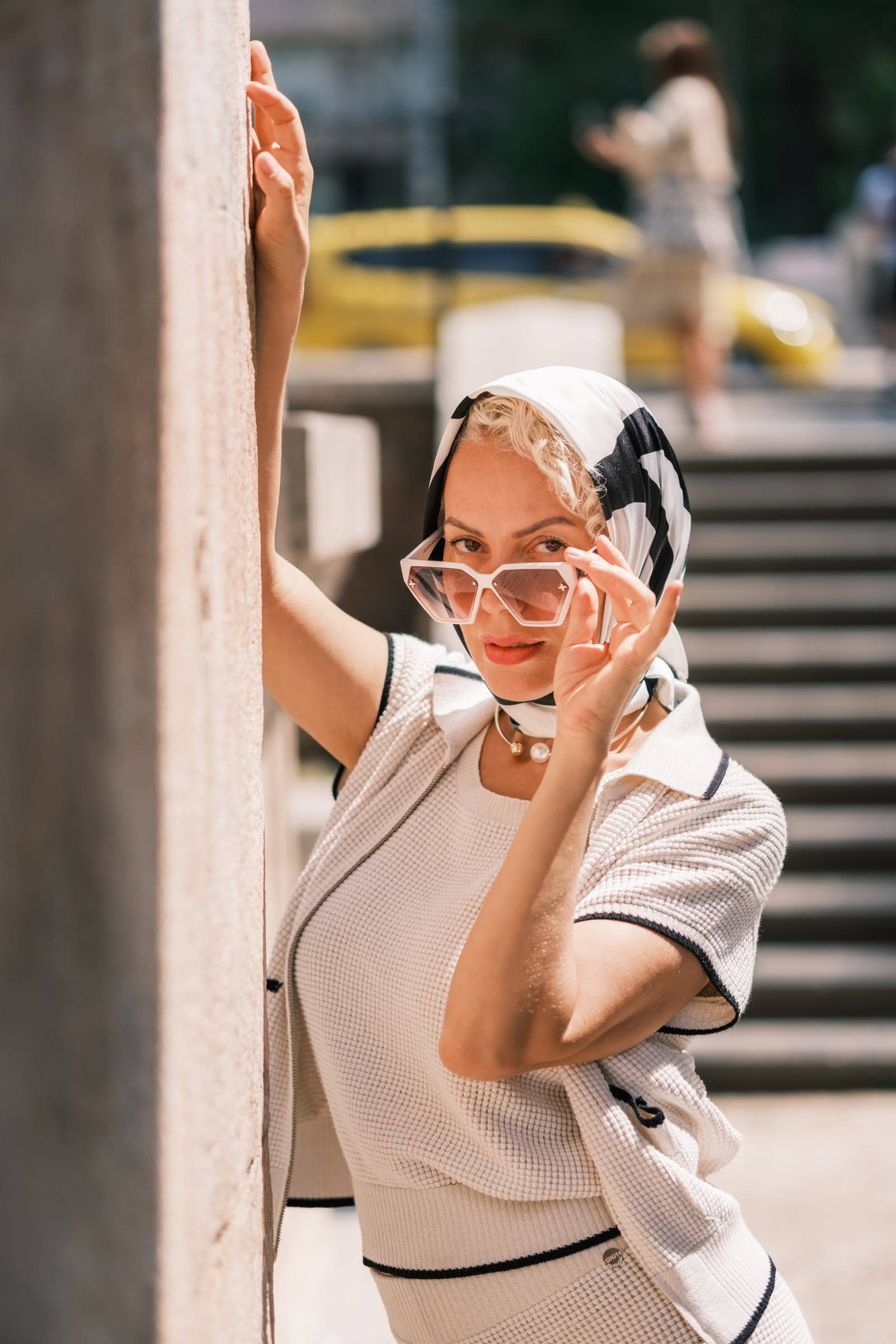 Woman in a silk scarf adjusting her sunglasses by a marble pillar